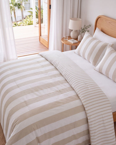 A neatly made bed in a bright bedroom with beige and white striped bedding, including a duvet cover and matching pillow shams, a wooden headboard, a bedside table with a lamp, and an open door leading to a wooden deck.