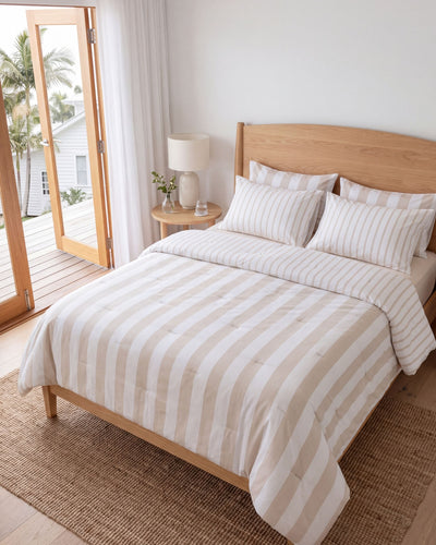 A neatly made bed in a bright bedroom with a light wood frame, striped cream and beige bedding, matching pillows, a wooden headboard, a small bedside table with a lamp, and an open door leading to a deck with palm trees.