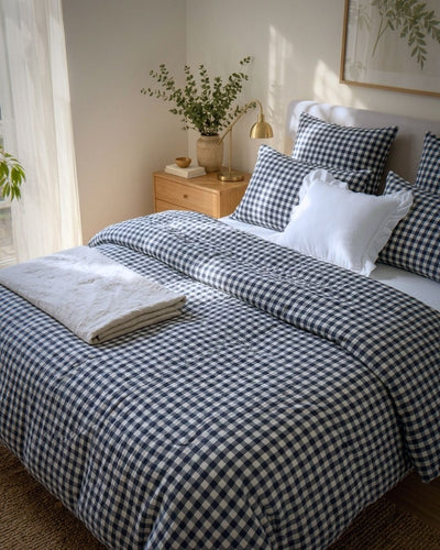 A neatly made bed in a softly lit bedroom, styled with a navy and white gingham checkered duvet cover, matching pillow shams, a white accent pillow, and a folded light gray throw blanket. A wooden nightstand, vase with greenery, and a gold floor lamp sit beside the bed.
