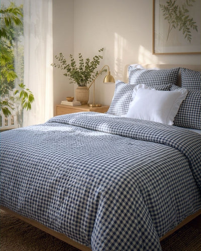 A neatly made bed in a softly lit bedroom, covered with a blue-and-white gingham check duvet and matching pillow shams, plus a few solid white pillows. A wooden nightstand, lamp, vase with greenery, and framed wall art are visible in the room.