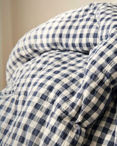A close-up of a soft, rumpled bedding item, likely a comforter or duvet, featuring a classic navy blue and off-white gingham check pattern.
