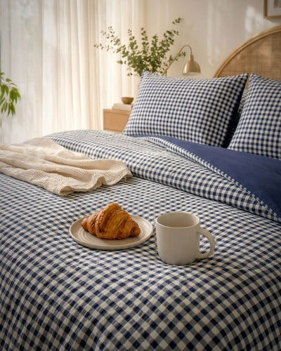 A neatly made bed with navy and white gingham bedding, including a duvet cover, pillowcases, and a solid navy throw blanket. A cream knit throw, a croissant on a small plate, and a white ceramic mug are placed on the bed in a softly lit bedroom setting.