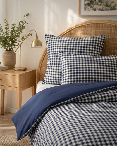 A neatly made bed with navy and white gingham check bedding, including two pillowcases and a duvet cover, styled in a warm bedroom setting with a wooden headboard, bedside table, lamp, and plant.