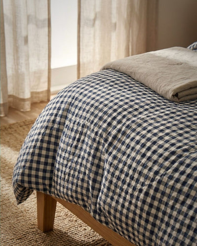 A navy and cream gingham checkered comforter on a wooden bed frame, styled with a folded beige throw blanket in a softly lit bedroom setting.