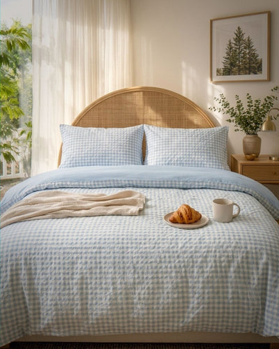 A neatly made bed in a softly lit bedroom, featuring a light blue and white gingham quilt, matching pillow shams, a woven rattan headboard, a cream throw blanket, a croissant on a plate, and a white mug on the bedside table.