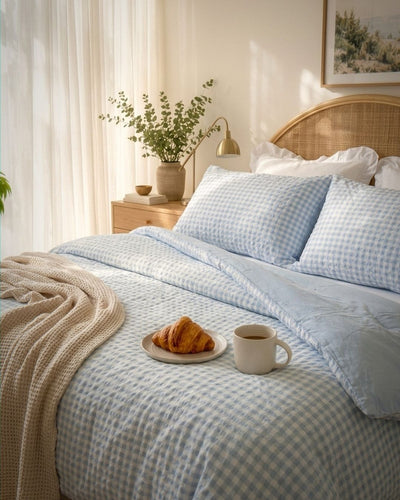 A neatly made bed in a softly lit bedroom, featuring light blue and white gingham bedding, matching pillows, a beige waffle-knit throw blanket, a wooden headboard, and a bedside table with a lamp and vase.
