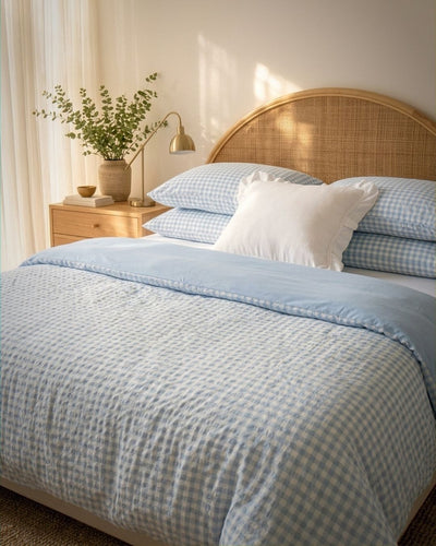 A neatly made bed in a softly lit bedroom, featuring a light blue and white gingham patterned duvet cover and matching pillows, with a solid white accent pillow. The bed has a round woven rattan headboard, and a wooden nightstand with a lamp and a vase of greenery sits on the left.