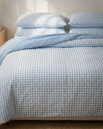 A neatly made bed with a light blue and white gingham checkered duvet cover and matching pillowcases, with a solid light blue folded blanket or sheet on top, photographed in a softly lit bedroom.