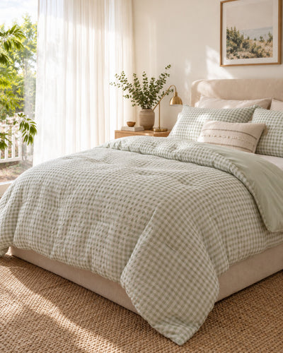 A neatly made bed in a bright bedroom, dressed with a light beige and sage green gingham comforter set, matching pillows, and a beige upholstered bed frame. Sunlight streams in from a window with sheer curtains, and a small bedside table with a lamp and plant is visible.
