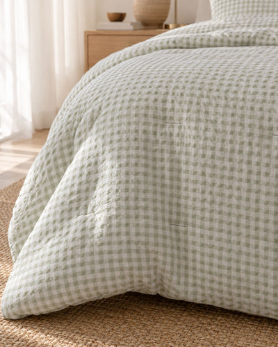 A close-up of a bed dressed in a light sage green and white gingham checkered quilt with a textured waffle weave, styled in a bright, sunlit bedroom.