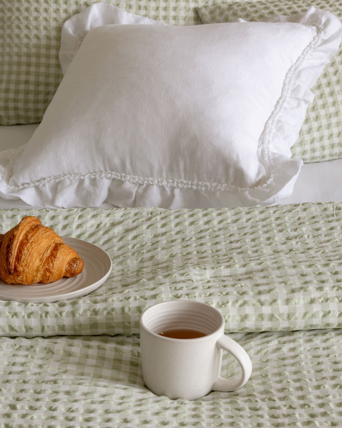 A white ruffled decorative pillow on a bed with a light green and white gingham check blanket and matching pillowcases, styled with a croissant on a plate and a white mug of tea in the foreground.