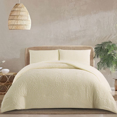 A modern bedroom features the Morgan and Reid Lemon Butter Gingham Comforter Set on a neatly made bed with matching pillowcases, rattan bedhead, neutral walls, woven pendant light, and a wooden side table with decorative accents.