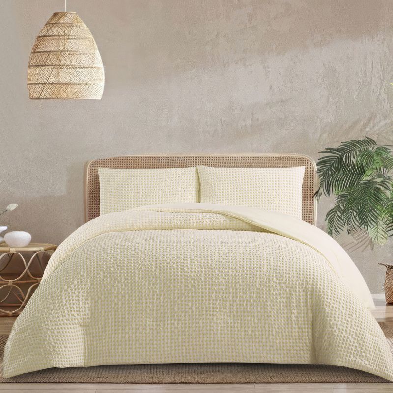 A modern bedroom features the Morgan and Reid Lemon Butter Gingham Comforter Set on a neatly made bed with matching pillowcases, rattan bedhead, neutral walls, woven pendant light, and a wooden side table with decorative accents.