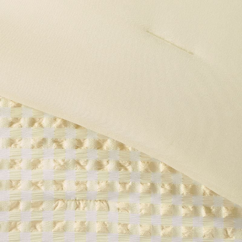 A modern bedroom features the Morgan and Reid Lemon Butter Gingham Comforter Set on a neatly made bed with matching pillowcases, rattan bedhead, neutral walls, woven pendant light, and a wooden side table with decorative accents.