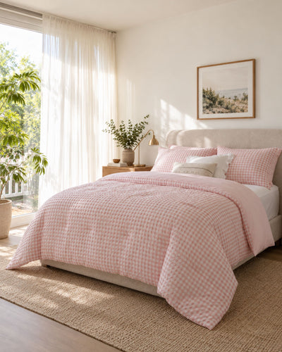 A neatly made bed in a bright bedroom featuring a light pink and white gingham checkered comforter and matching pillows, styled with a beige upholstered headboard, a woven rug, and soft natural light from a window.