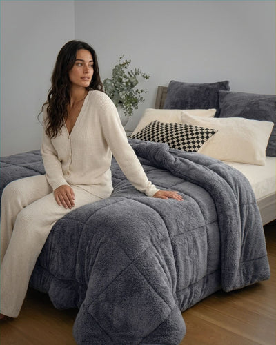 Woman sitting on a bed with a gray comforter in a cozy bedroom setting.