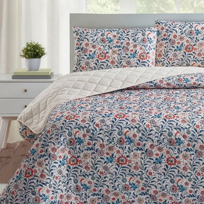 A bright bedroom features the Morgan and Reid Indian Floral Coverlet Set with matching pillow shams, complemented by white walls, large windows, a small lamp, plant, and alarm clock on bedside tables.