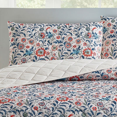 A bright bedroom features the Morgan and Reid Indian Floral Coverlet Set with matching pillow shams, complemented by white walls, large windows, a small lamp, plant, and alarm clock on bedside tables.