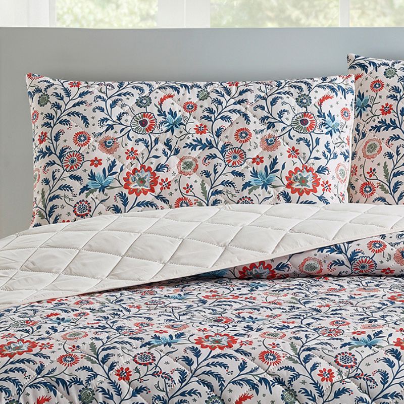 A bright bedroom features the Morgan and Reid Indian Floral Coverlet Set with matching pillow shams, complemented by white walls, large windows, a small lamp, plant, and alarm clock on bedside tables.