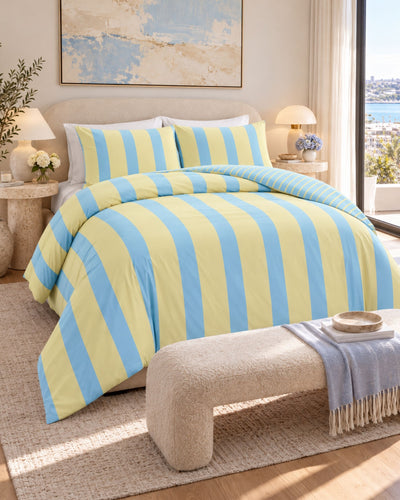 A bed dressed in a yellow and light blue striped bedding set, styled in a bright, modern bedroom with a bench, lamps, and a window overlooking the sea.