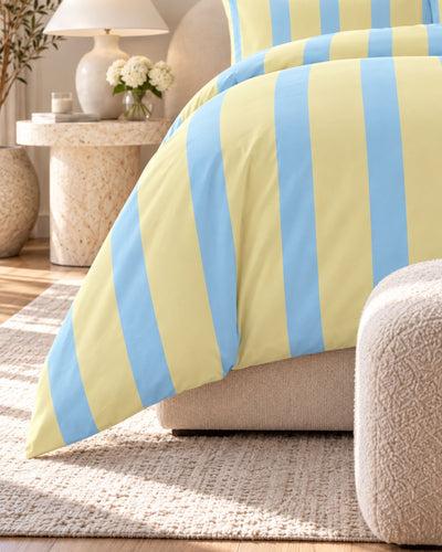 A close-up of a striped bedding set in pale yellow and light blue, styled on a bed in a warm, sunlit bedroom with a textured rug, side table, and lamp.