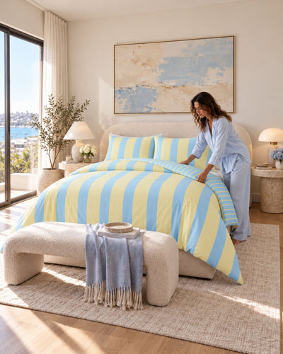 A woman in light blue pajamas adjusts a yellow and blue striped duvet cover on a bed in a bright, modern bedroom. The room features a matching blue and yellow striped bedding set, a textured cream bench, and a large abstract painting on the wall.