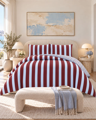 A bed dressed in a red, white, and blue striped bedding set, styled in a bright, modern bedroom with a beige upholstered bench, bedside tables, lamps, and a large abstract painting on the wall.