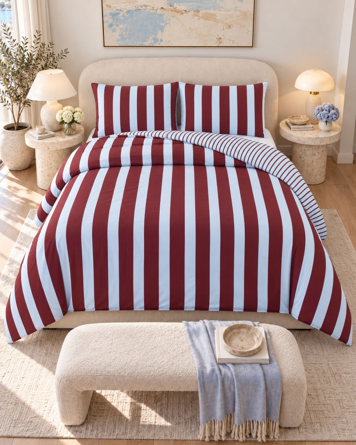 A bed dressed in a red and white vertically striped duvet cover and matching pillow shams, styled in a bright, modern bedroom with a bench at the foot, bedside tables, lamps, and a neutral wall art piece.