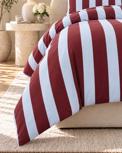 A close-up of a bed dressed in a burgundy and white vertically striped duvet cover and pillowcase, styled in a warm, sunlit bedroom with a beige rug and side table.