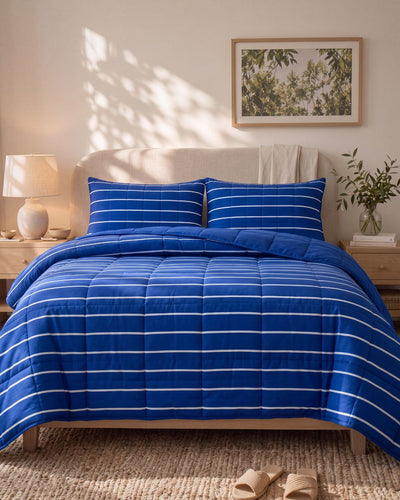 A blue quilted comforter with white horizontal stripes and matching striped pillowcases, styled on a bed in a warm, sunlit bedroom with bedside tables, lamps, and wall art.