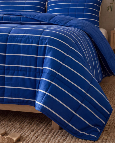 A blue quilted comforter with white horizontal stripes, styled on a bed with matching pillows in a bedroom setting.