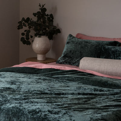 Bed with green velvet bedding and pink pillows, vase with plants on nightstand.
