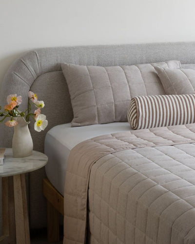 Neatly made bed with beige bedding and striped pillow, side table with flowers in a room.