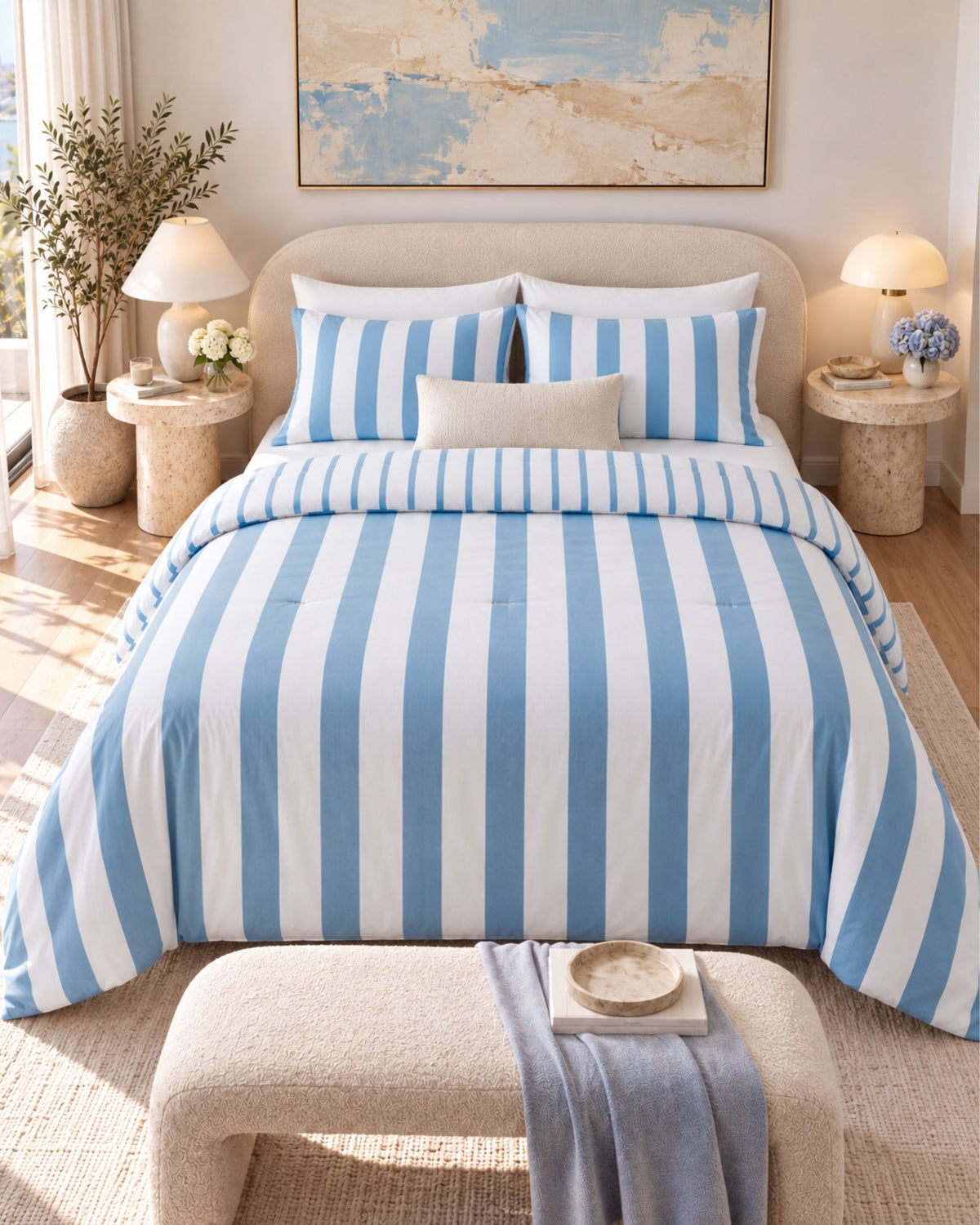 A neatly made bed in a bright bedroom setting, featuring a blue and white vertically striped comforter, matching striped pillow shams, two white pillows, and a small light beige accent pillow. The room includes a beige upholstered headboard, bedside tables with lit lamps, a bench at the foot of the bed, and framed wall art.
