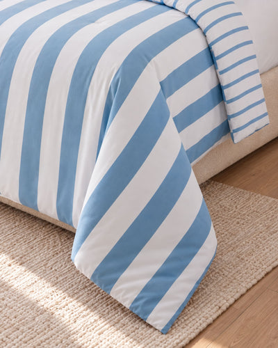 A striped bedding comforter in light blue and white, draped over the side of a bed on a woven beige rug over a light wood floor.