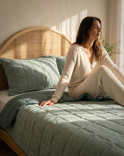 Woman sitting on a bed with green bedding and a wicker headboard.