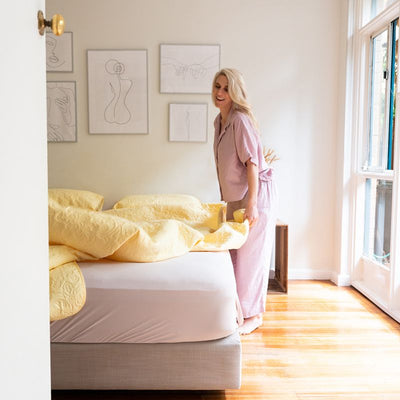The Morgan and Reid Serene Butter Coverlet Set by Morgan and Reid, featuring a yellow coverlet and two matching pillows, brightens the room. A wooden nightstand with dried flowers and minimalist line art complete the cozy look.