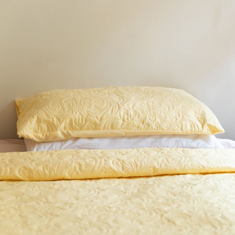 The Morgan and Reid Serene Butter Coverlet Set by Morgan and Reid, featuring a yellow coverlet and two matching pillows, brightens the room. A wooden nightstand with dried flowers and minimalist line art complete the cozy look.