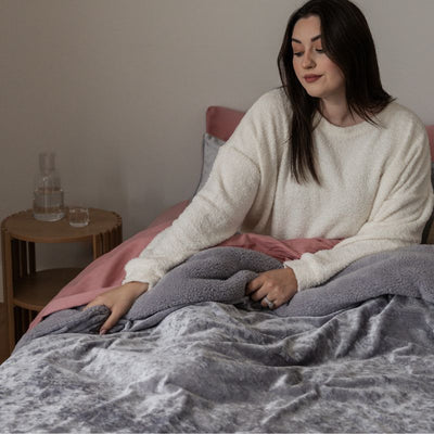 Woman sitting on a bed wrapped in a gray blanket, with a side table and water pitcher in the background.