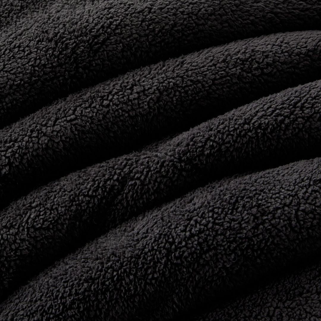 A modern bedroom features a neatly made bed with the Morgan and Reid Black Snuggle Fleece Comforter Set, accented by a white pillow, a side table with a plant and books, wall art, a pendant lamp, and sheer curtains letting in natural light.