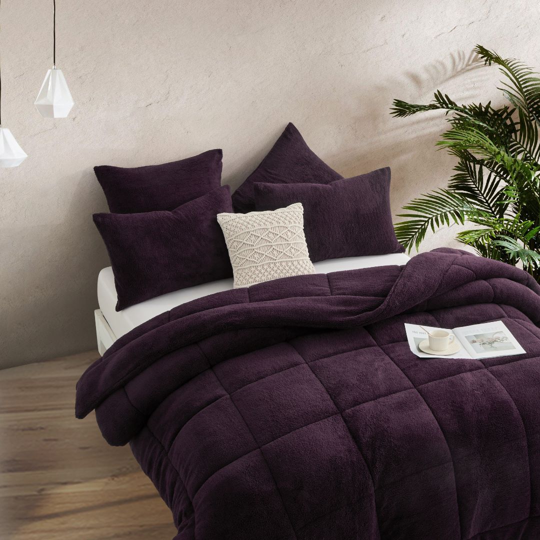 A neatly made bed with the Morgan and Reid Plum Snuggle Fleece Comforter Set and pillows sits on a light rug. Pendant lights hang above, with a side table, vase, and a large potted plant against a textured beige wall.