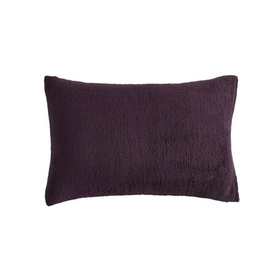 A neatly made bed with the Morgan and Reid Plum Snuggle Fleece Comforter Set and pillows sits on a light rug. Pendant lights hang above, with a side table, vase, and a large potted plant against a textured beige wall.