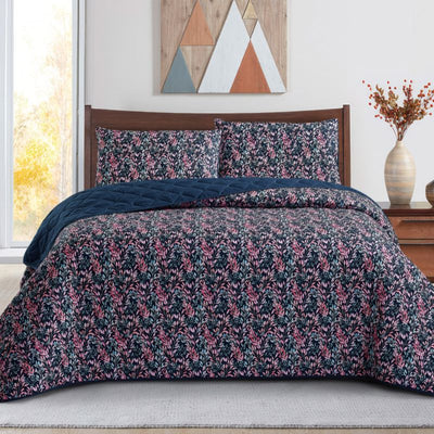 A neatly made bed with a dark floral comforter and matching pillows, the Morgan and Reid Sophia Coverlet Set in lightweight breathable fabric at the foot, a wooden headboard, geometric wall art, and a vase of flowers on the bedside table.