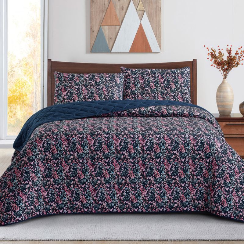 A neatly made bed with a dark floral comforter and matching pillows, the Morgan and Reid Sophia Coverlet Set in lightweight breathable fabric at the foot, a wooden headboard, geometric wall art, and a vase of flowers on the bedside table.