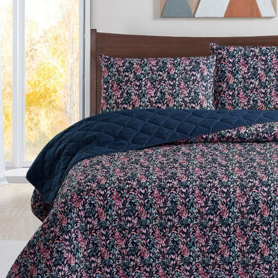 A neatly made bed with a dark floral comforter and matching pillows, the Morgan and Reid Sophia Coverlet Set in lightweight breathable fabric at the foot, a wooden headboard, geometric wall art, and a vase of flowers on the bedside table.
