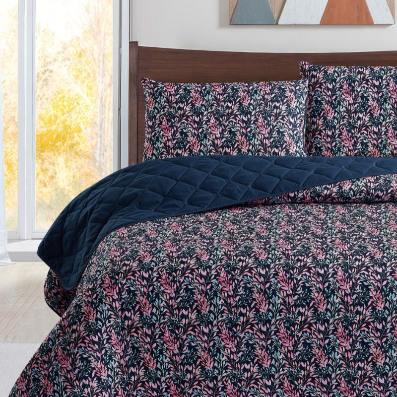 A neatly made bed with a dark floral comforter and matching pillows, the Morgan and Reid Sophia Coverlet Set in lightweight breathable fabric at the foot, a wooden headboard, geometric wall art, and a vase of flowers on the bedside table.