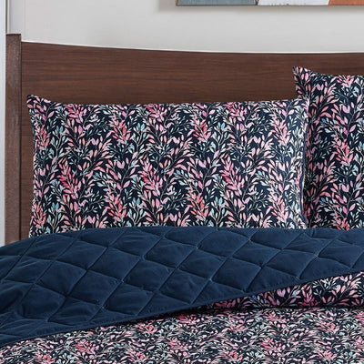 A neatly made bed with a dark floral comforter and matching pillows, the Morgan and Reid Sophia Coverlet Set in lightweight breathable fabric at the foot, a wooden headboard, geometric wall art, and a vase of flowers on the bedside table.
