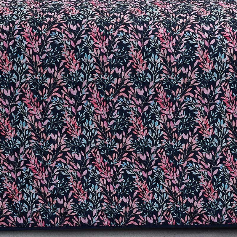 A neatly made bed with a dark floral comforter and matching pillows, the Morgan and Reid Sophia Coverlet Set in lightweight breathable fabric at the foot, a wooden headboard, geometric wall art, and a vase of flowers on the bedside table.