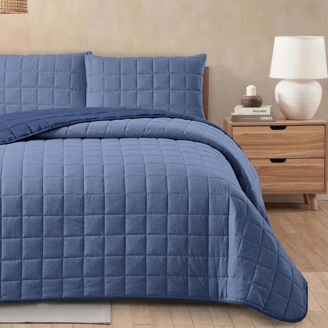 Blue quilted bedding set on a bed with a wooden nightstand and lamp in the background.
