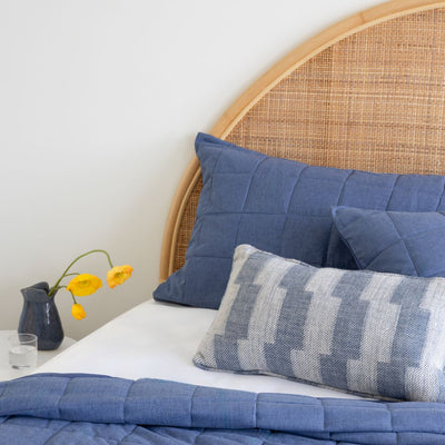 Bed with blue bedding and pillows, wicker headboard, and small vase with yellow flowers on a white surface.